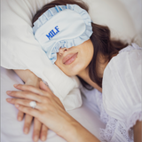A woman lying in bed with her eyes covered by a light blue sleep mask with the text 'MLF' in blue. She is wearing a white lace-trimmed nightgown and resting on white pillows.