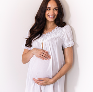 A smiling pregnant woman wearing a white short-sleeve maternity nightgown with lace trim, standing against a plain white wall.