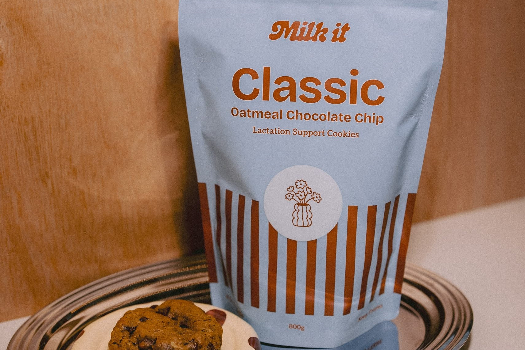 Milk it cookie dough bundle: blue Classic Oatmeal Chocolate Chip pouch on a tray beside a baked cookie on a spotted dish