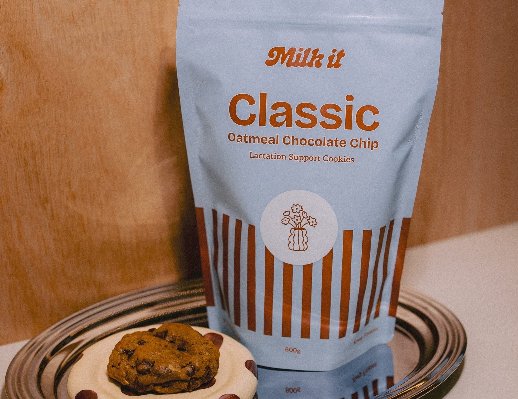 Milk it cookie dough bundle: blue Classic Oatmeal Chocolate Chip pouch on a tray beside a baked cookie on a spotted dish