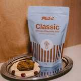 Milk it cookie dough bundle: blue Classic Oatmeal Chocolate Chip pouch on a tray beside a baked cookie on a spotted dish