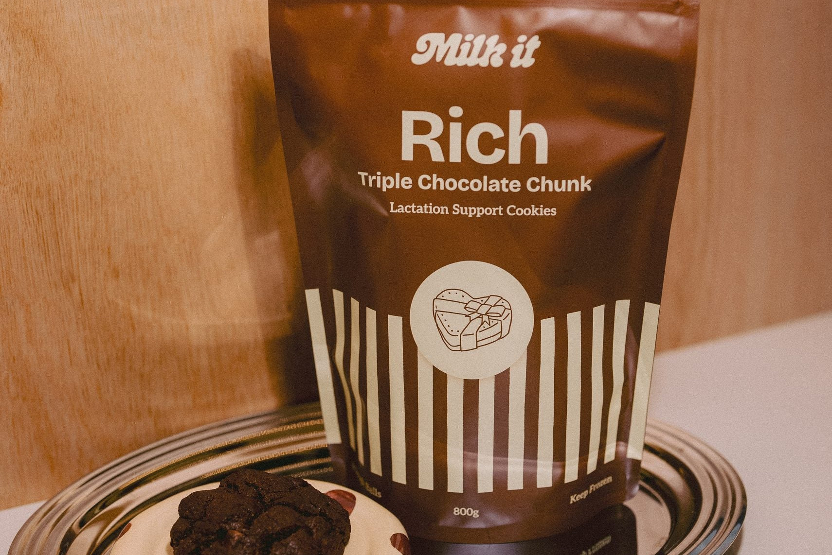 Milk it cookie dough bundle: brown Rich Triple Chocolate Chunk pouch on a tray beside a dark baked cookie on a spotted dish