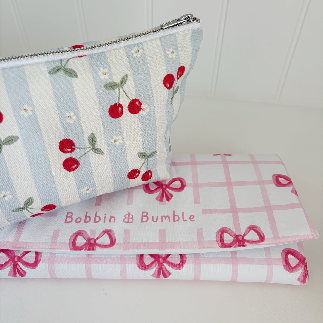 Nappy Wallet Cherry Stripe, angled view beside a folded pink bow gingham changing mat with the Bobbin and Bumble name