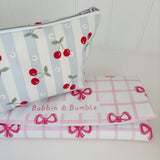 Nappy Wallet Cherry Stripe, angled view beside a folded pink bow gingham changing mat with the Bobbin and Bumble name