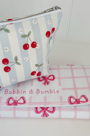 Nappy Wallet Cherry Stripe, angled view beside a folded pink bow gingham changing mat with the Bobbin and Bumble name