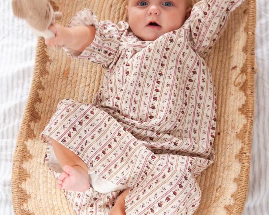 Beatrice Rose Stripe baby nightie, baby lying in a woven basket with arms raised, holding a soft toy