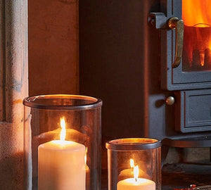 Palm Beach Hurricane Lanterns in two sizes holding lit pillar candles on a hearth beside a log burner