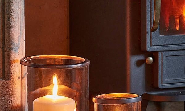Palm Beach Hurricane Lanterns in two sizes holding lit pillar candles on a hearth beside a log burner
