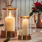 Palm Beach Hurricane Lantern in two sizes with glass cylinders and metal bases, each holding a lit pillar candle on a shelf