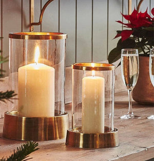 Palm Beach Hurricane Lantern in two sizes with glass cylinders and metal bases, each holding a lit pillar candle on a shelf
