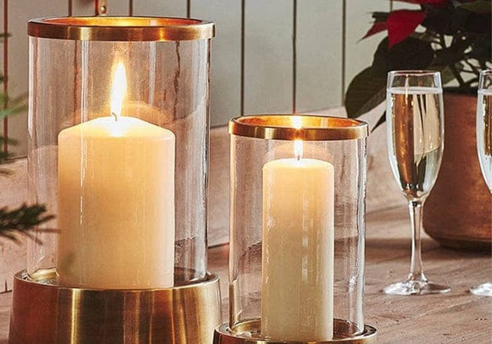 Palm Beach Hurricane Lantern in two sizes with glass cylinders and metal bases, each holding a lit pillar candle on a shelf