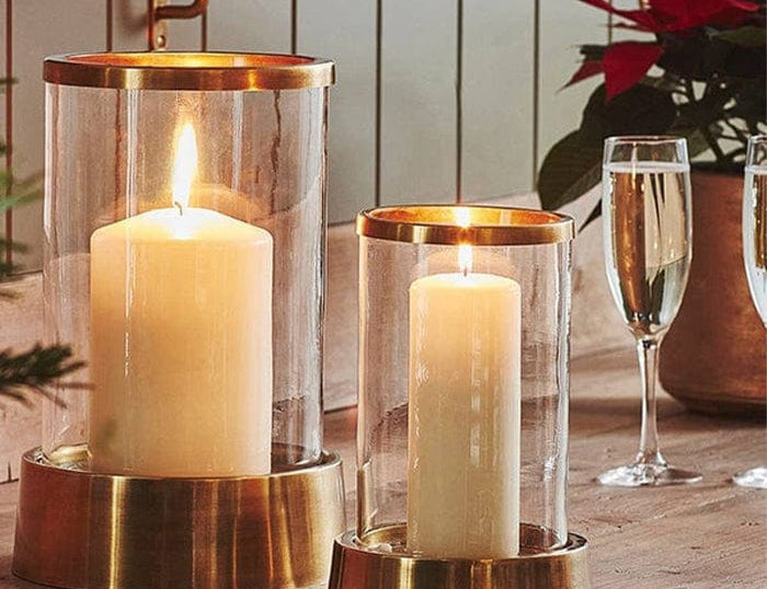 Palm Beach Hurricane Lantern in two sizes with glass cylinders and metal bases, each holding a lit pillar candle on a shelf