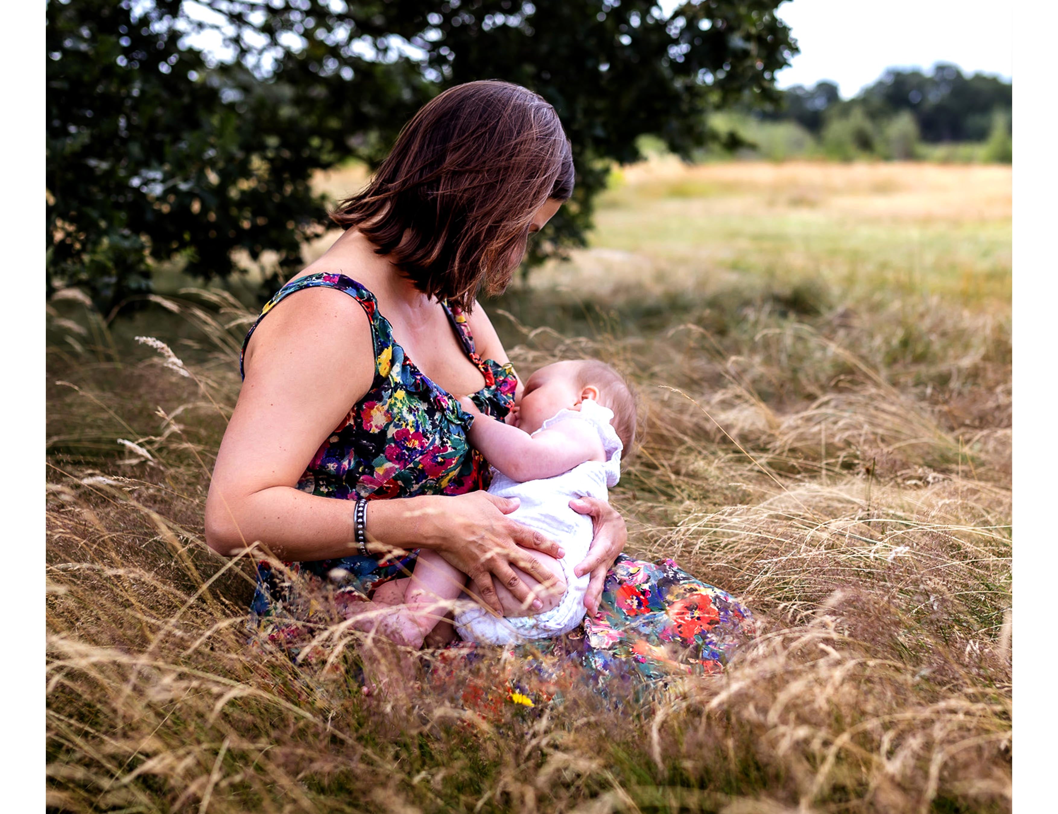 A woman in a colorful floral maternity dress breastfeeding a baby in a grassy outdoor field under a tree. The text 'Annie Breastfeeding in the Dali dress' is written at the bottom.