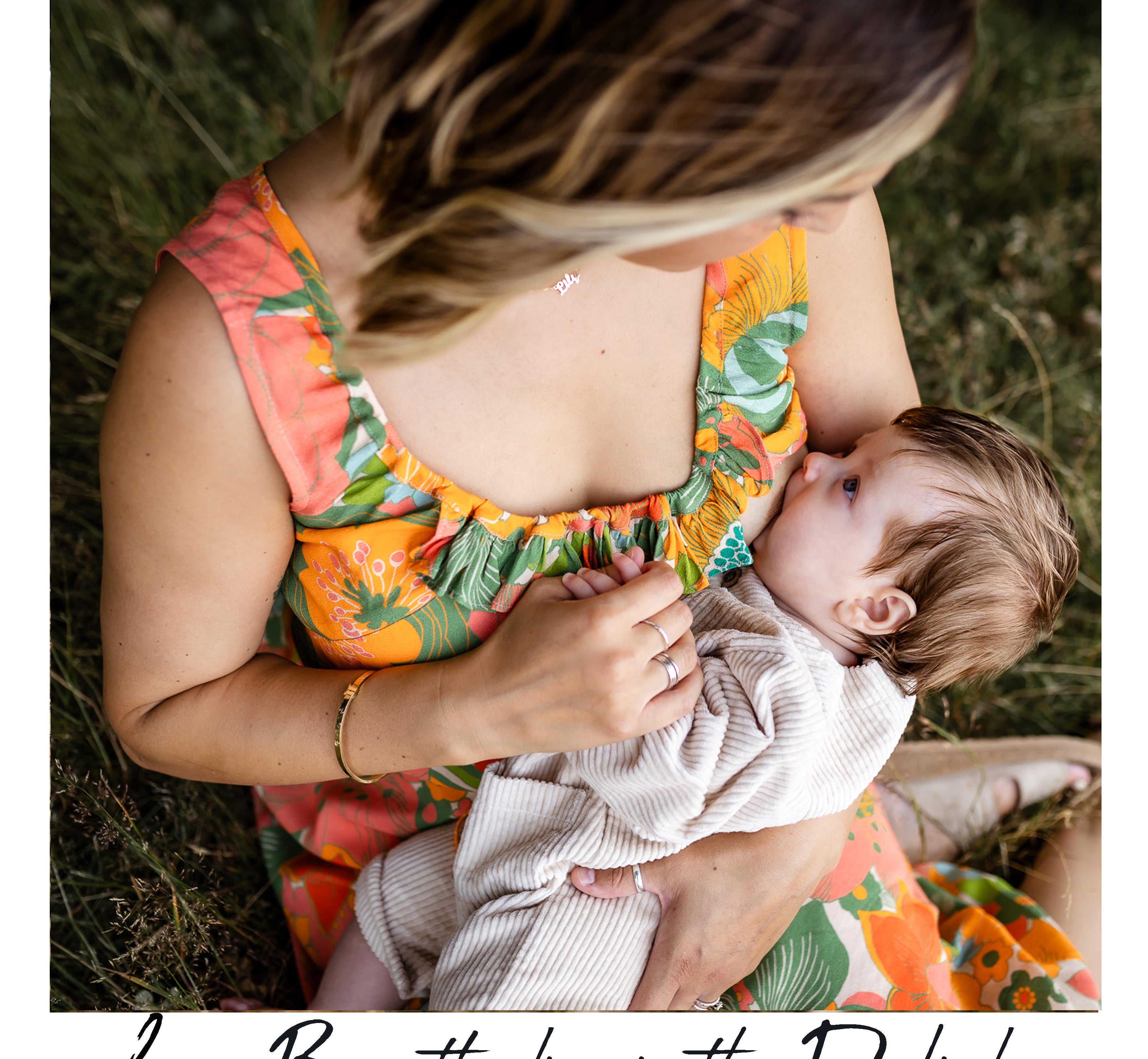 A woman in a colorful floral Dali dress breastfeeding a baby outdoors on grass. The photo has a white border with the text 'Lucy Breastfeeding in the Dali dress' at the bottom.
