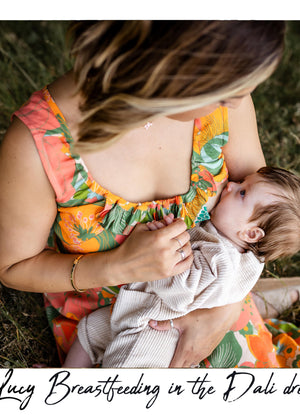 A woman in a colorful floral Dali dress breastfeeding a baby outdoors on grass. The photo has a white border with the text 'Lucy Breastfeeding in the Dali dress' at the bottom.