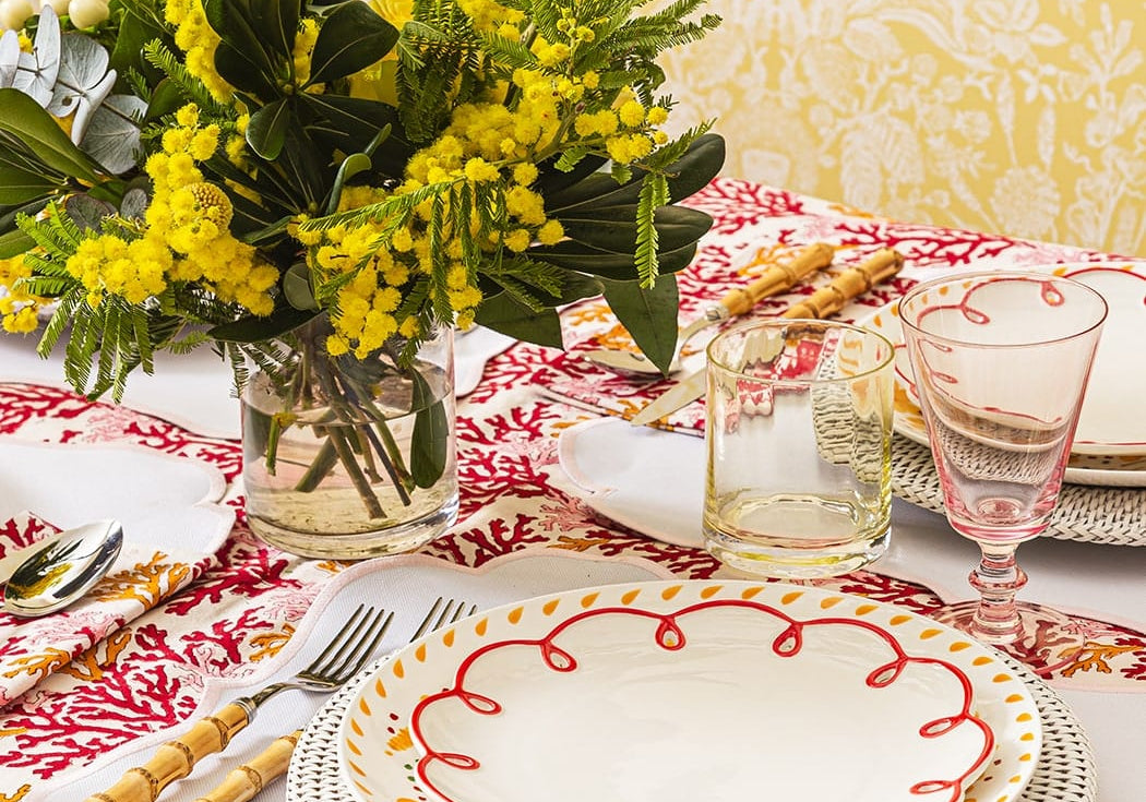 Rosy Red Scalloped Lunch Plate on a woven charger on a table with mimosa flowers, bamboo-handled cutlery and a pink glass