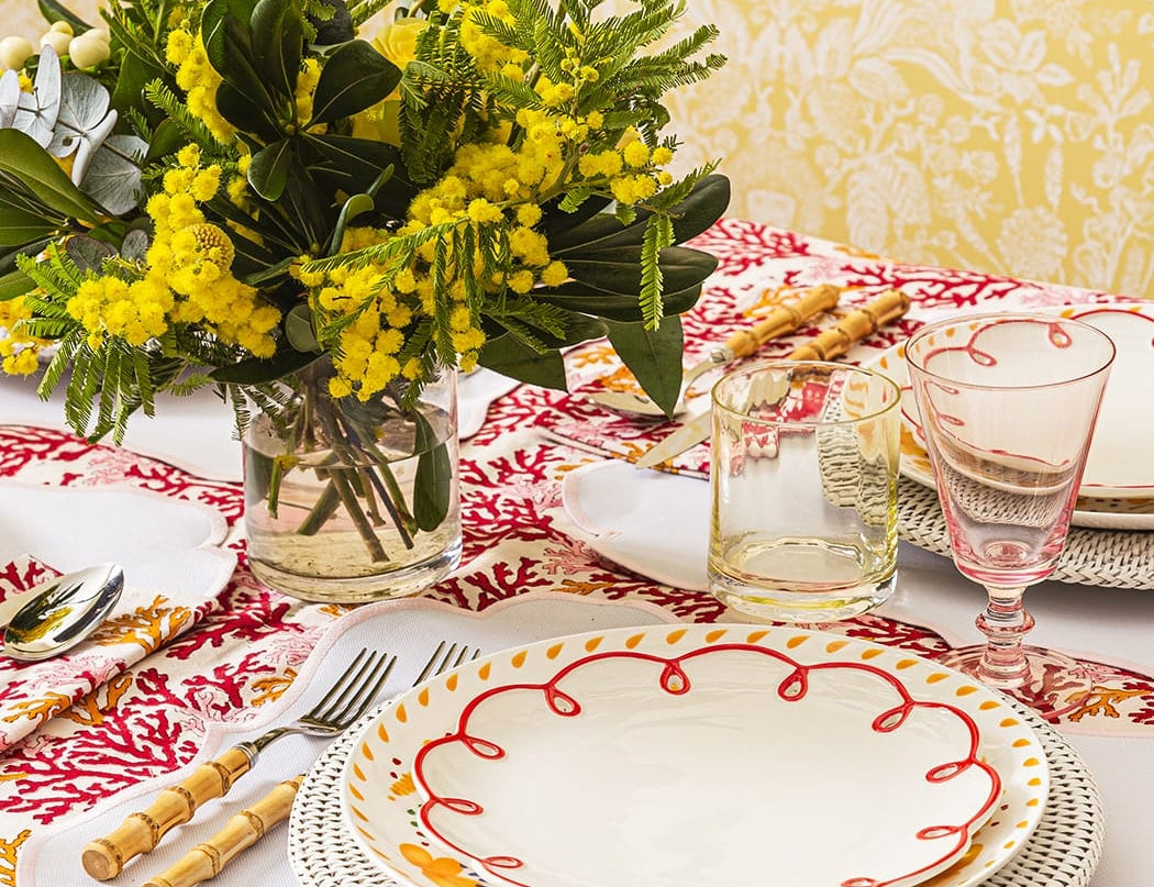 Rosy Red Scalloped Lunch Plate on a woven charger on a table with mimosa flowers, bamboo-handled cutlery and a pink glass