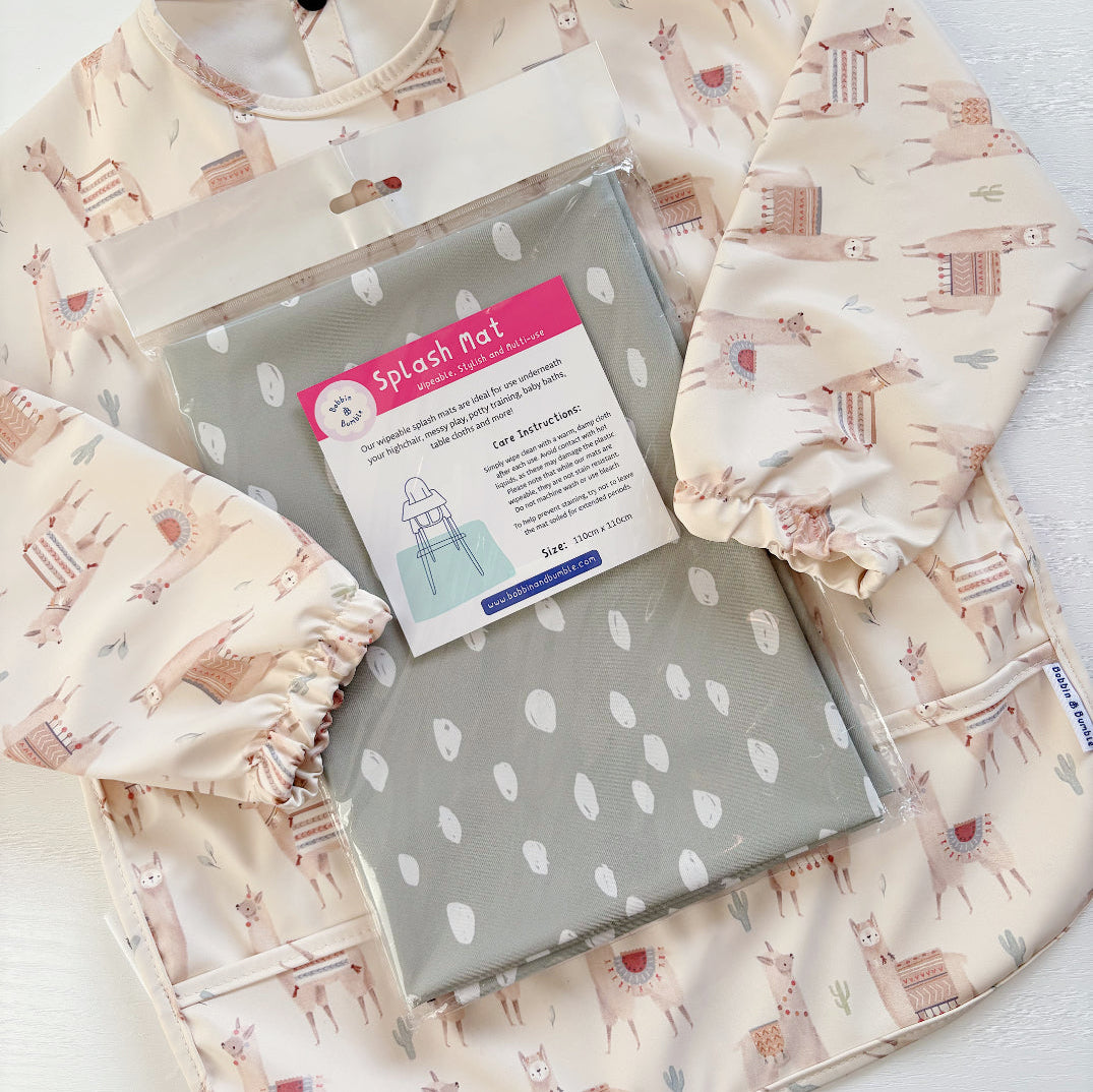 Splash Mat Sage Spots, in its packaging resting on a cream llama-print long-sleeved bib