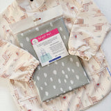Splash Mat Sage Spots, in its packaging resting on a cream llama-print long-sleeved bib