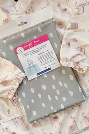 Splash Mat Sage Spots, in its packaging resting on a cream llama-print long-sleeved bib