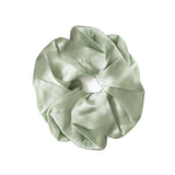 A sage green satin hair scrunchie with a gathered, pleated texture and a white elastic center band, shown on a plain white background.