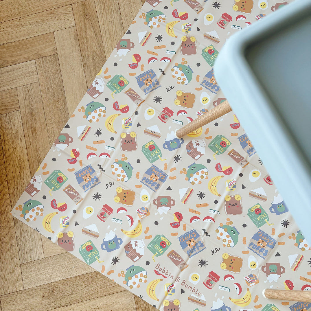 Splash Mat Snack Time seen from above on a herringbone wood floor, with highchair legs and tray edge at the side