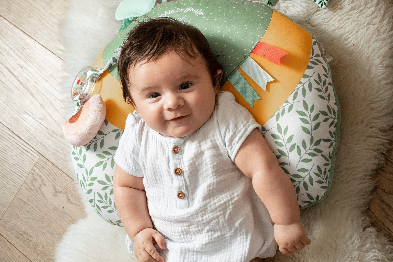 Sophie la girafe Cosy Play Cushion, seen from above with a smiling baby lying on it on a sheepskin rug