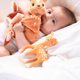 FanFan the Fawn Comforter, orange, held to the mouth by a baby lying on white bedding beside a fawn toy