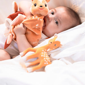 FanFan the Fawn Comforter, orange, held to the mouth by a baby lying on white bedding beside a fawn toy