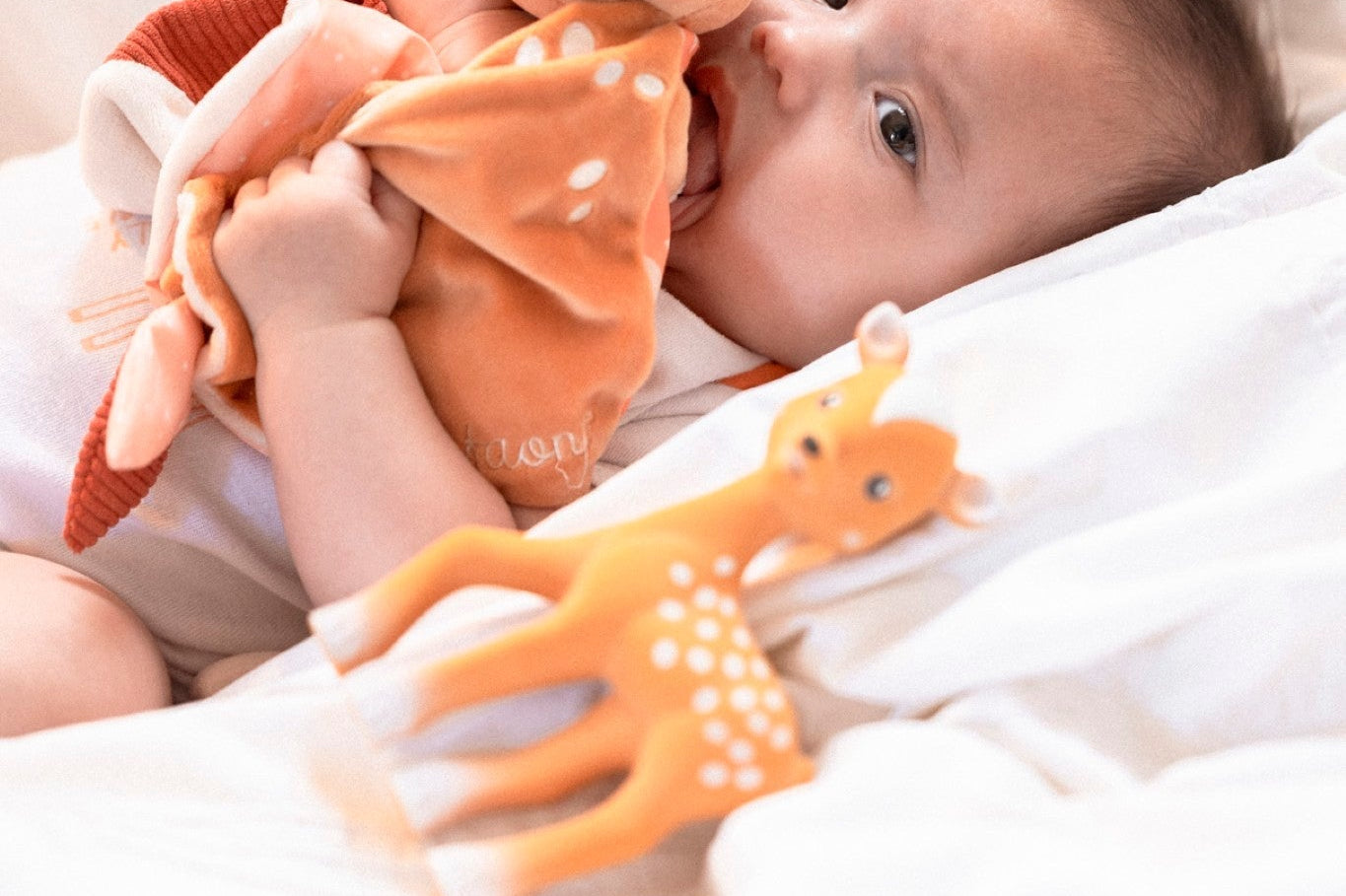 FanFan the Fawn Comforter, orange, held to the mouth by a baby lying on white bedding beside a fawn toy