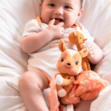 FanFan the Fawn Comforter, orange, held on the lap of a smiling baby in a white bodysuit with a fawn toy