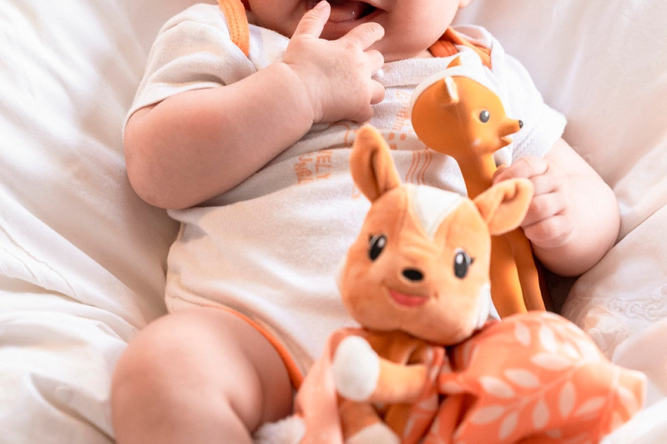 FanFan the Fawn Comforter, orange, held on the lap of a smiling baby in a white bodysuit with a fawn toy