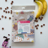 Splash Mat Snack Time folded in its clear packaging with a label, beside bananas and chocolate cereal rings
