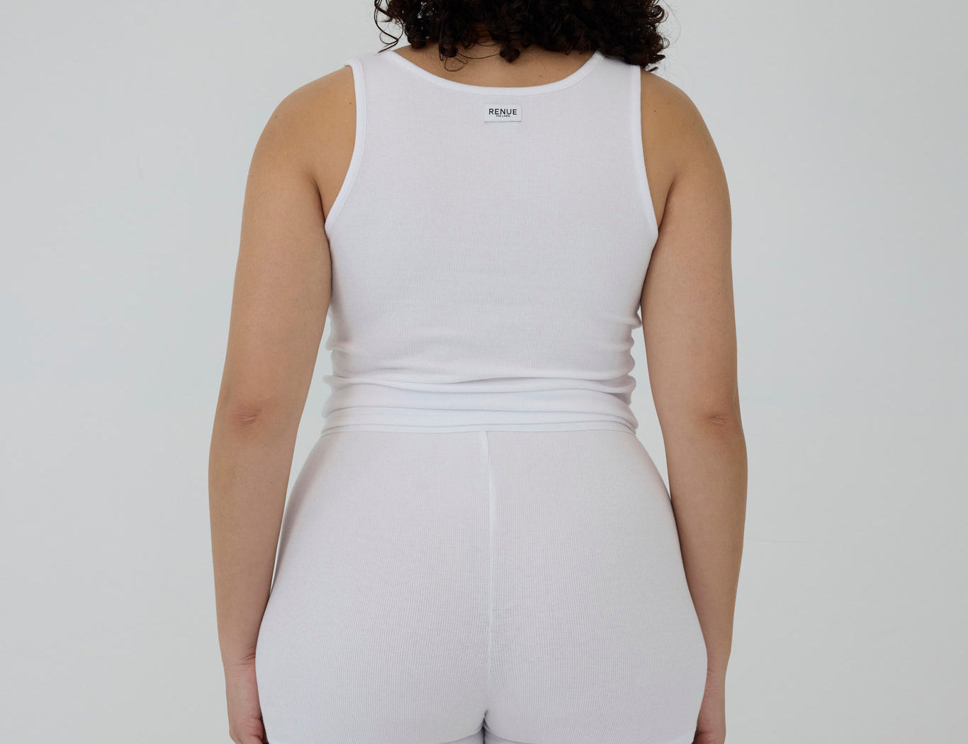 A person shown from the back wearing a white sleeveless tank top and matching white shorts against a plain light gray studio background.
