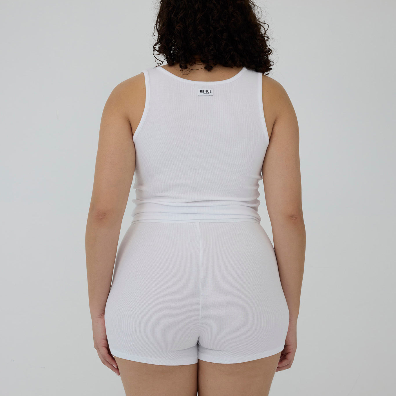 A person shown from the back wearing a white sleeveless tank top and matching white shorts against a plain light gray studio background.