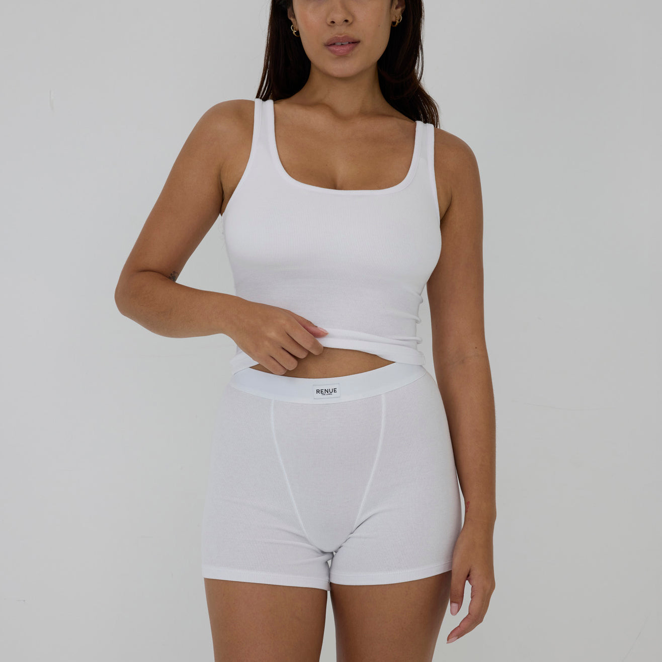 A woman modeling a white ribbed tank top and matching white boxer briefs against a plain light gray studio background.