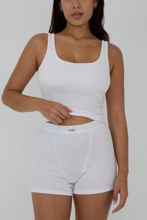 A woman modeling a white ribbed tank top and matching white boxer briefs against a plain light gray studio background.