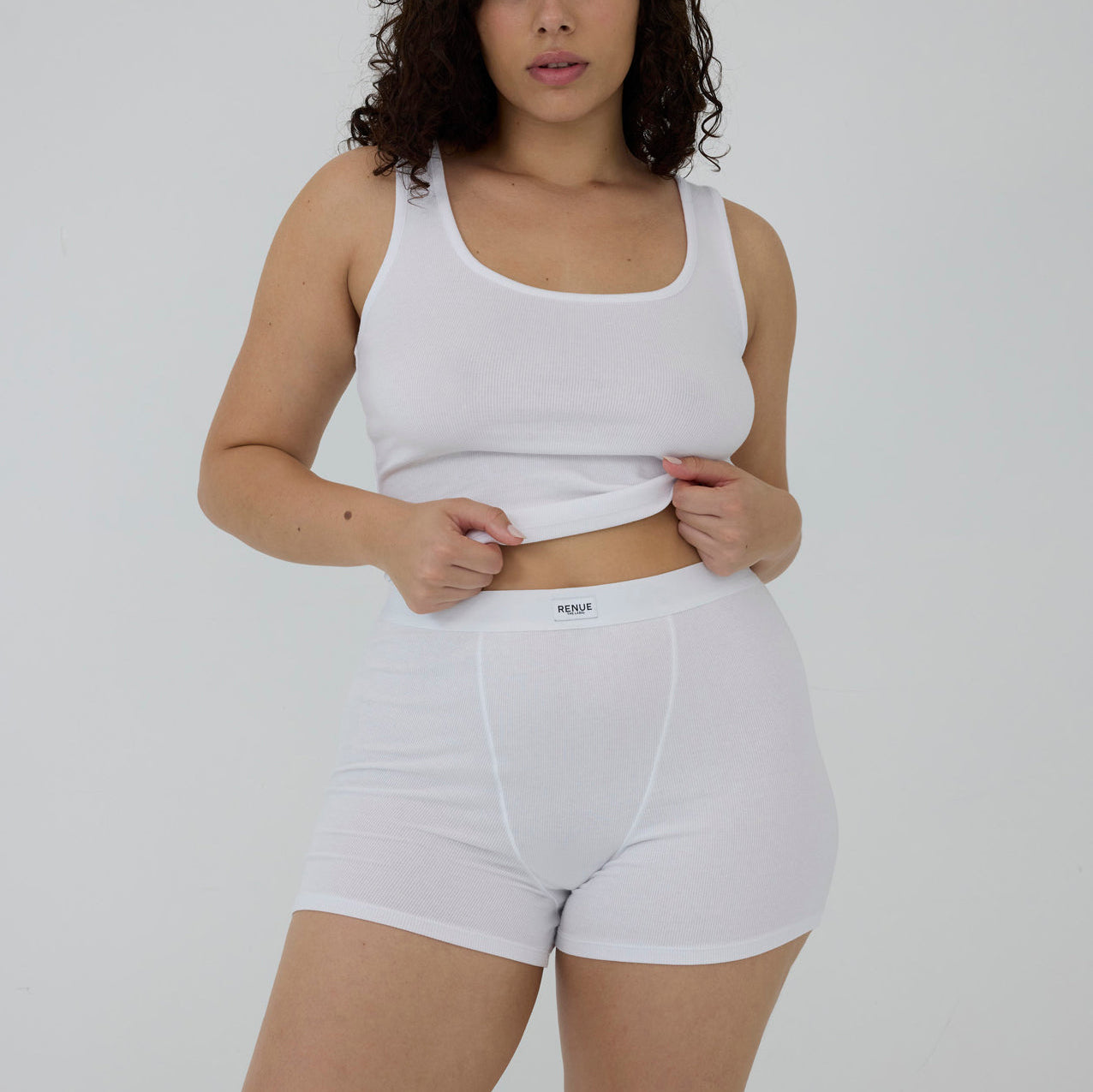 A model wearing a white ribbed tank top and matching white briefs, lifting the waistband of the briefs to show the brand label 'MEDI'.