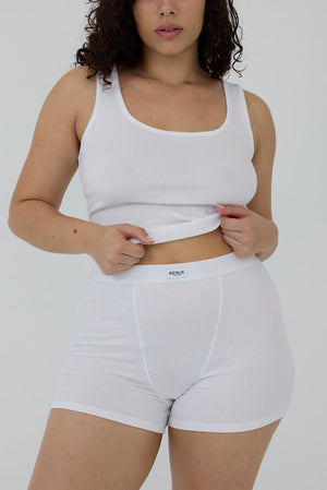 A model wearing a white ribbed tank top and matching white briefs, lifting the waistband of the briefs to show the brand label 'MEDI'.