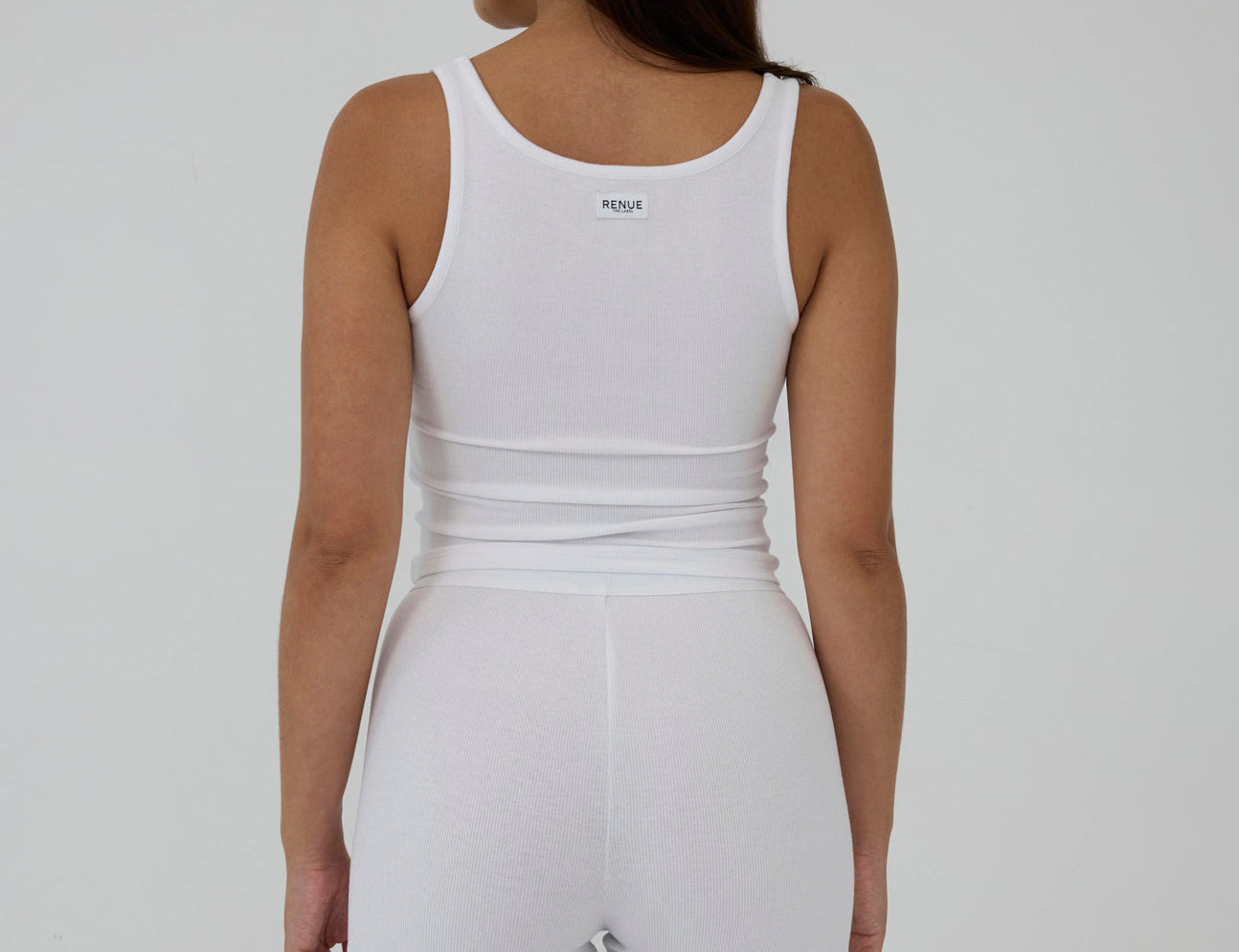 A woman shown from the back wearing a white sleeveless tank top and matching white shorts against a plain light gray studio background.