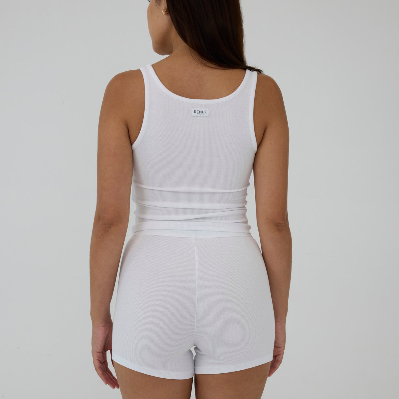 A woman shown from the back wearing a white sleeveless tank top and matching white shorts against a plain light gray studio background.